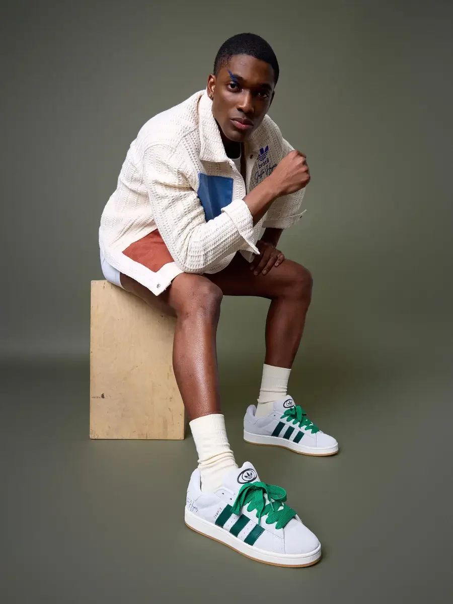 JUS10H x Adidas Brasil campaign, model seated on wood block pulling jacket collar, white textured patchwork jacket with blue and terracotta suede panels, Adidas Campus 00s in white with dark green stripes and green laces foregrounded, sage green back