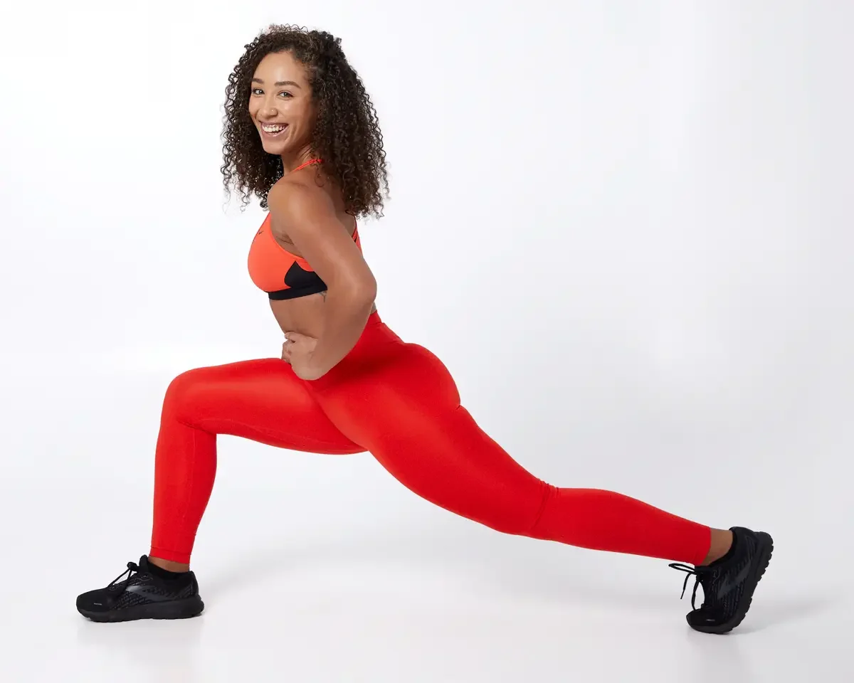 Model portfolio development session, commercial athletic look, orange sports bra with black underband, high-waist orange leggings, black athletic sneakers, full-length, deep lunge profile, smiling over shoulder, white background
