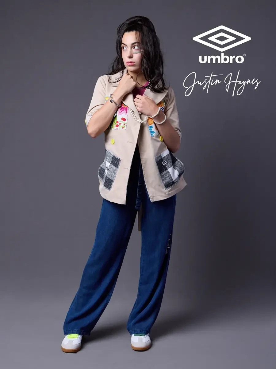 Editorial fashion photography for the Umbro France collection. Vanessa in a three-quarter stance holding her collar and glancing sideways in a sand utility jacket with floral patchwork panels over a fuchsia t-shirt and wide-leg dark indigo jeans, sty