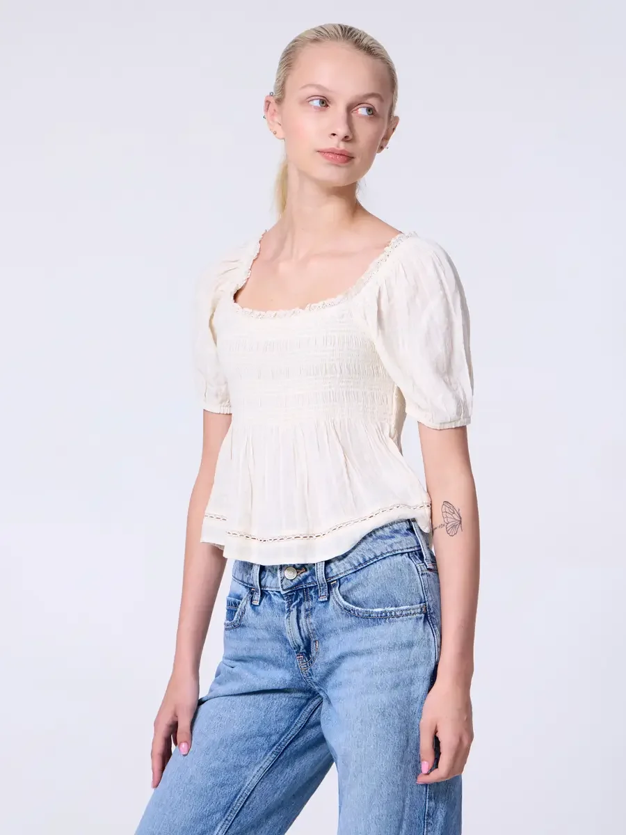 Model portfolio development session, commercial look, cream smocked square-neck puff-sleeve crop top with lace-trimmed neckline and crochet hem detail, three-quarter, slight turn, gaze directed off-camera, smocking texture and puff sleeve shown in cl