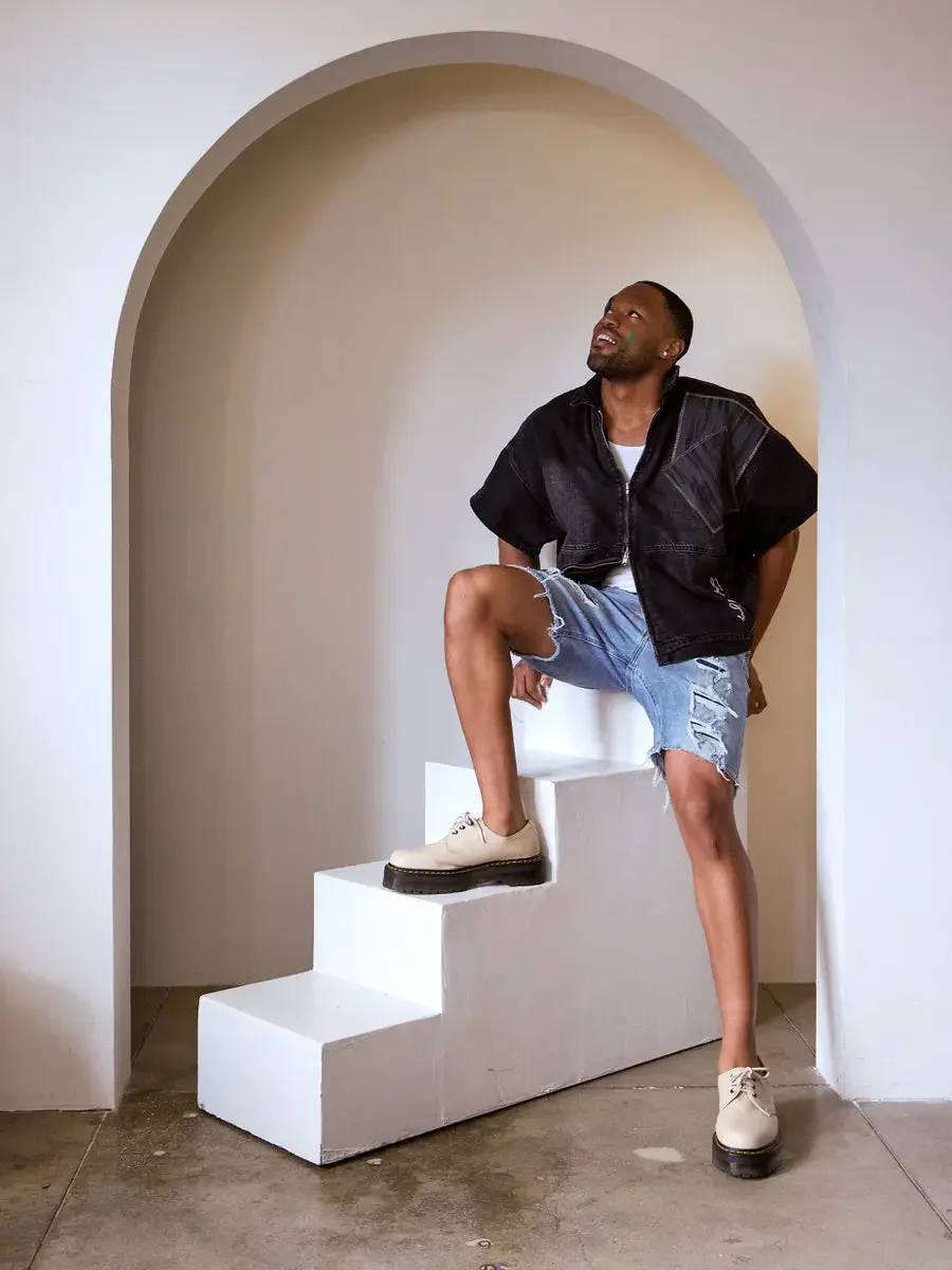 JUS10H Black Summer Jacket content library, male model in black denim wide-shoulder jacket with gray patchwork panels, foot up on white steps inside arch, looking up, distressed denim shorts, tan lace-up shoes, warm neutral background, photographed b