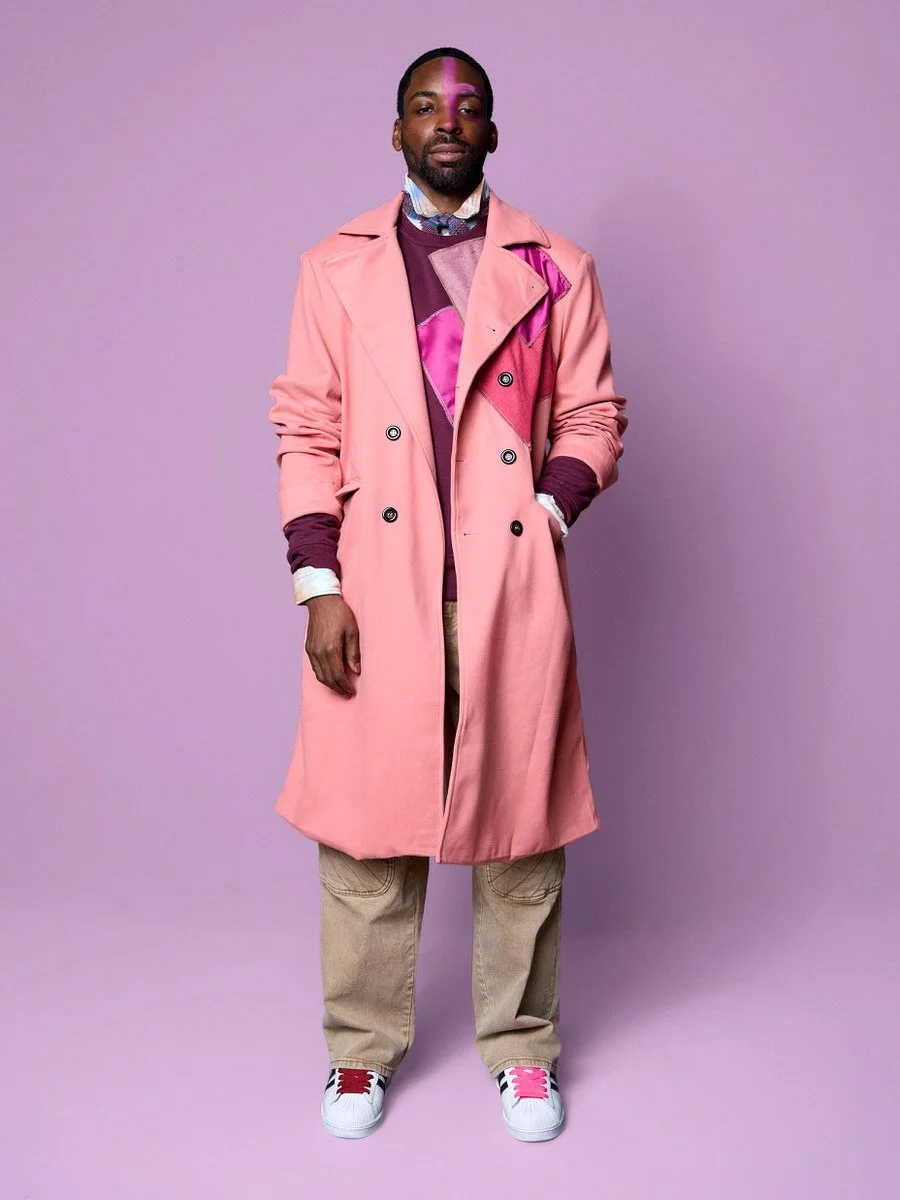 A man with dark skin and short hair stands against a lavender background, wearing a pink trench coat over a colorful sweater and beige pants, with white sneakers.