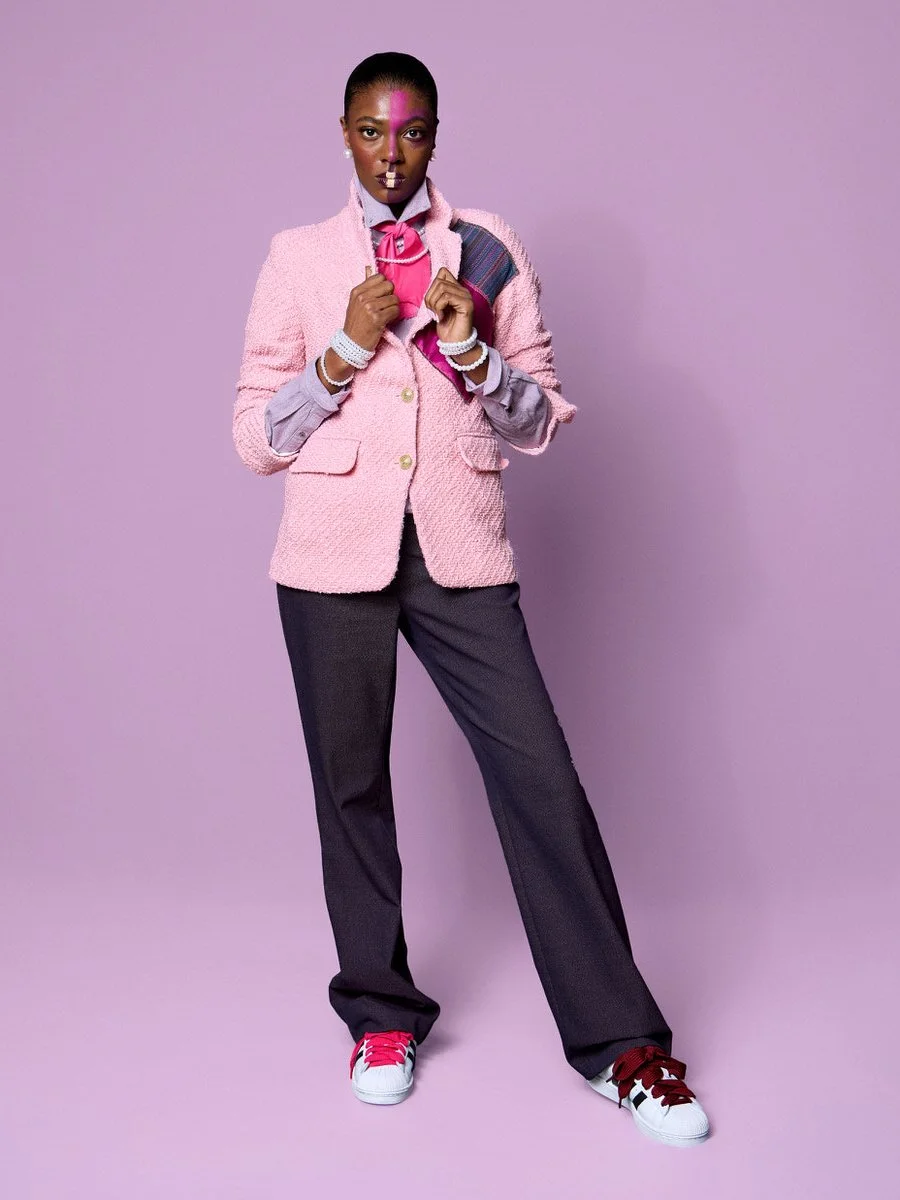 A woman standing against a purple background wearing a pink textured blazer, gray pants, pink and white sneakers, and jewelry, with face makeup and a serious expression.