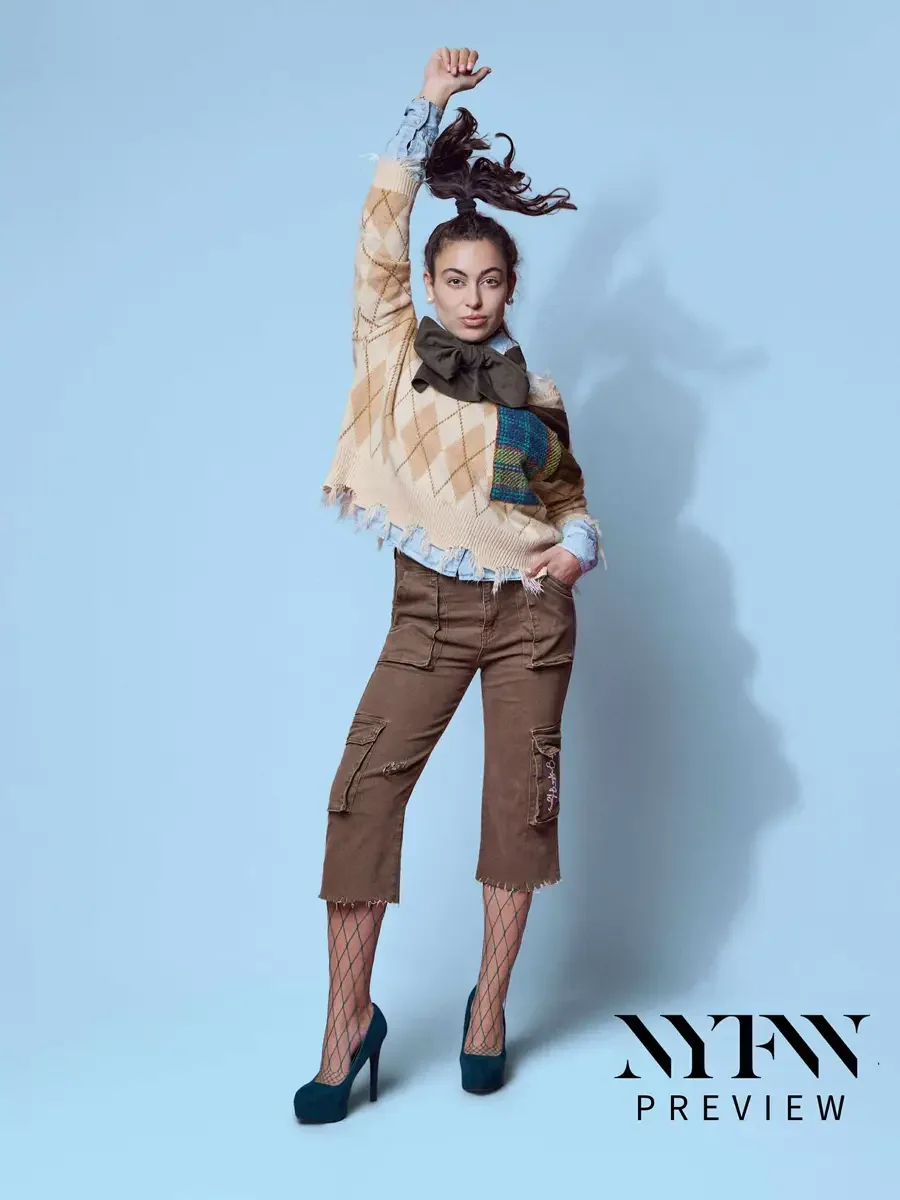 JUS10H 2025 promotional image, model in motion with arm raised, camel argyle fringe sweater with multicolor tartan patchwork panel, olive oversized bow, and brown distressed cargo capris over fishnet tights and teal platform heels, light blue backgro