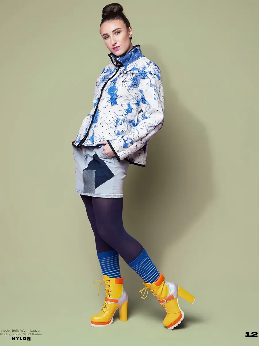 JUS10H 2025 Nylon Japan feature, model in white and blue geometric print zip-front jacket with JUS10H script detail over light denim patchwork mini skirt, navy tights, blue striped socks and yellow and orange platform lace-up boots, sage green backgr