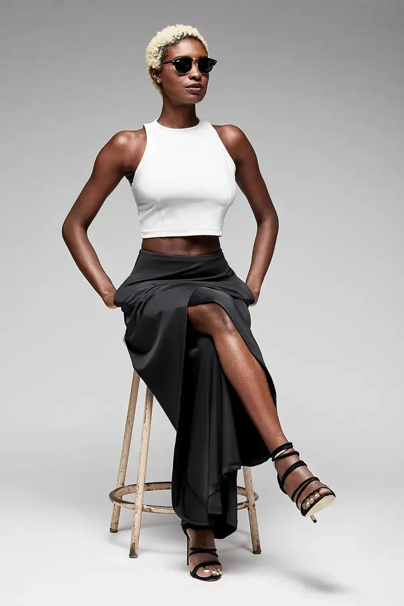 Minika Ko lookbook, black wide-leg trousers with asymmetric wrap skirt overlay and white halter crop top, seated view showing drape and movement of skirt overlay panel, dark sunglasses, photographed by Scott Parker Photo