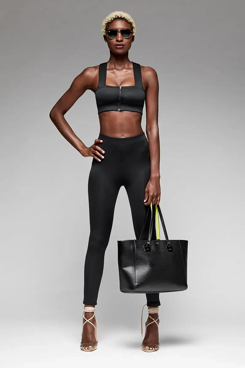 Minika Ko lookbook, black zip-front crop top and high-waist legging set, three-quarter view, center front zip closure, black structured tote with neon yellow strap detail, dark sunglasses, photographed by Scott Parker Photo