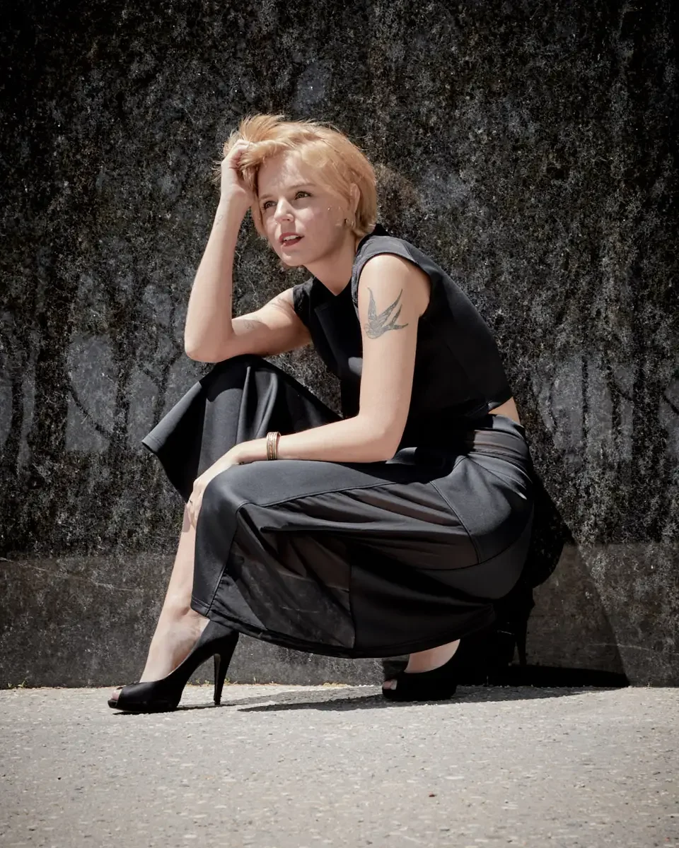 Satine, full length, crouching against granite wall, hand at temple, black structured cap-sleeve crop top, charcoal grey A-line midi skirt with sheer mesh panel, black satin peep-toe stiletto heels, gold bangles, bird tattoo on right upper arm visibl