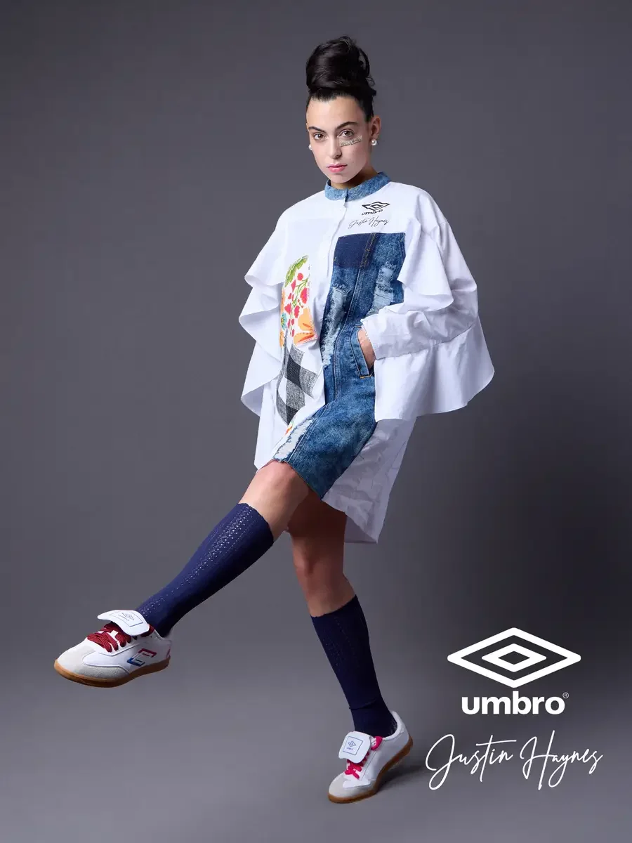 Fashion brand photography for the Umbro France collection. Vanessa in a side-step stance with one knee raised in an oversized white shirt dress with dramatic ruffled sleeves, a distressed denim center panel, and floral patchwork, styled by Justin Hay