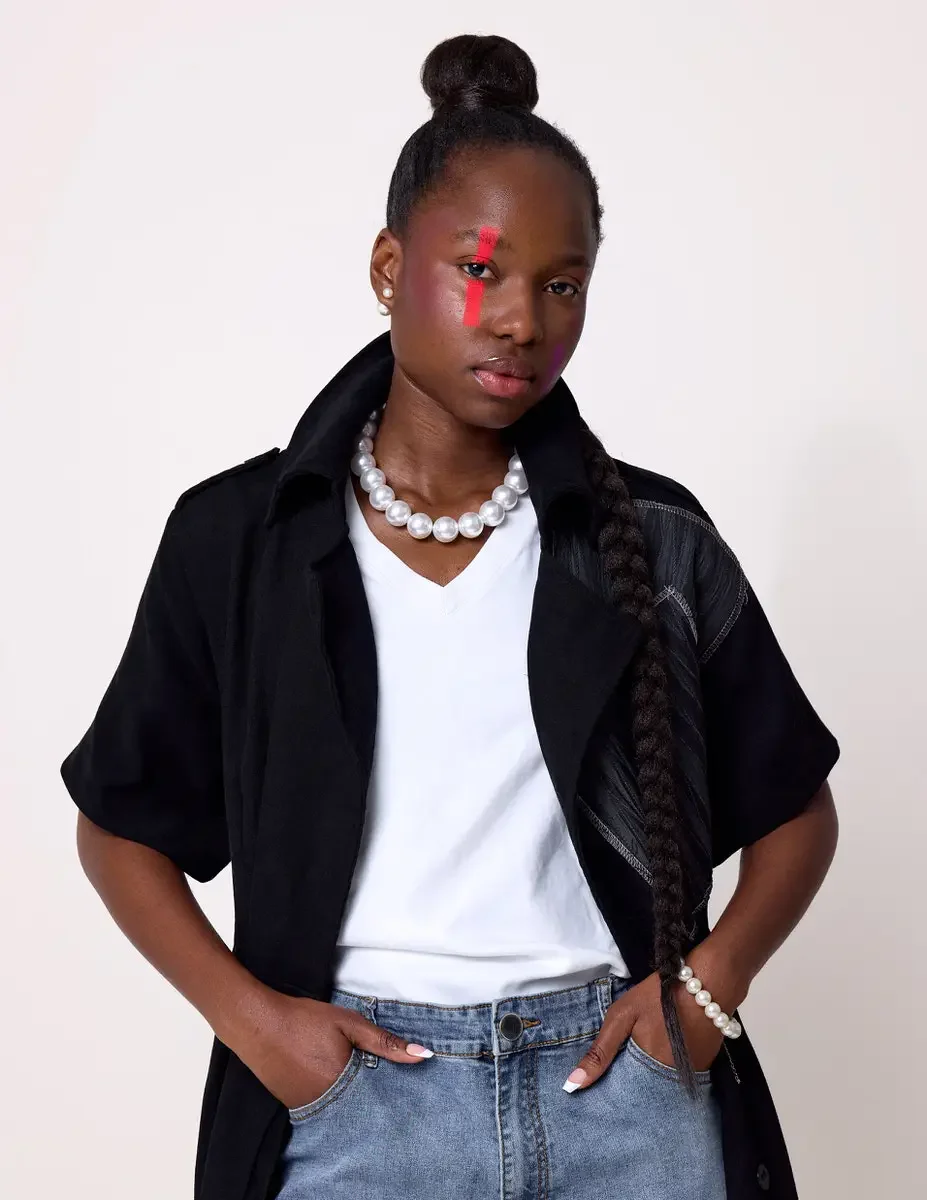 JUS10H Black Summer Jacket editorial, close portrait of female model in black long-line short-sleeve jacket with gray patchwork panel worn open, pearl necklace and bracelet, white tee, light-wash jeans, white background, photographed by Scott Parker 