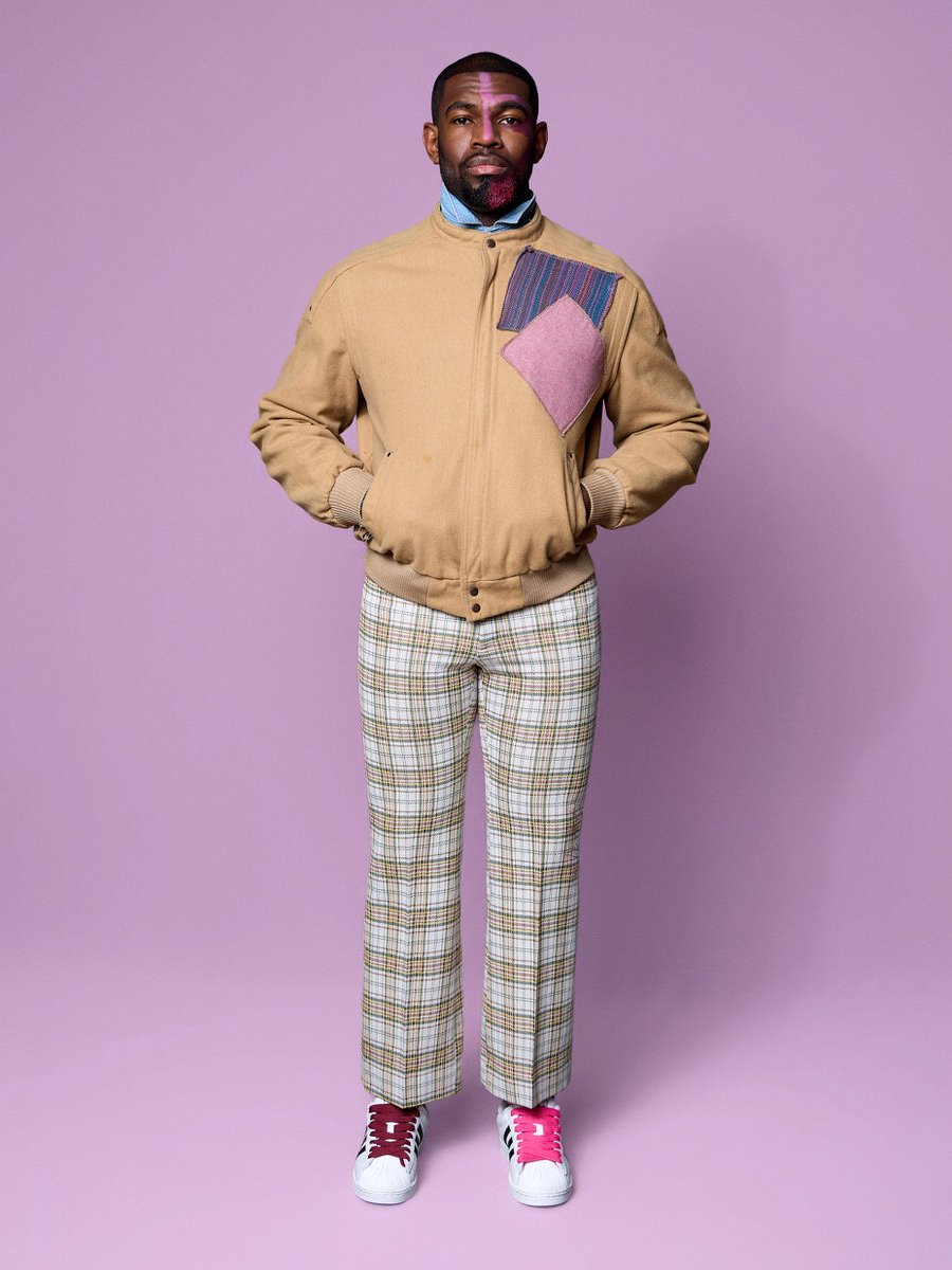 A man stands with hands in pockets against a purple background, wearing a beige jacket with a multi-colored patch on the chest, checkered pants, and white sneakers with red and pink accents.