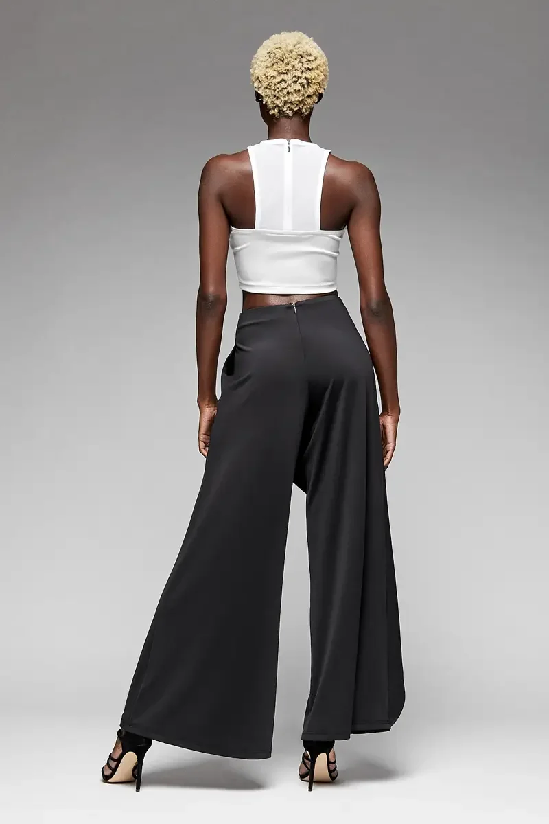 Minika Ko lookbook, black wide-leg trousers with asymmetric wrap skirt overlay and white halter crop top, back view, center back zip on trousers, draped skirt panel visible at side, racerback crop top with center back zip, photographed by Scott Parke