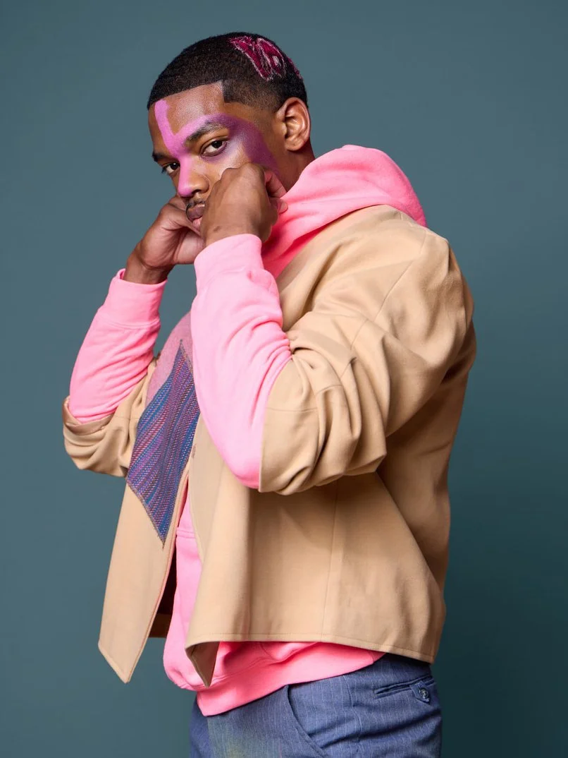 A young man with styled hair and pink face paint, wearing a beige jacket with pink sleeves and a pink hoodie underneath, poses against a plain background.