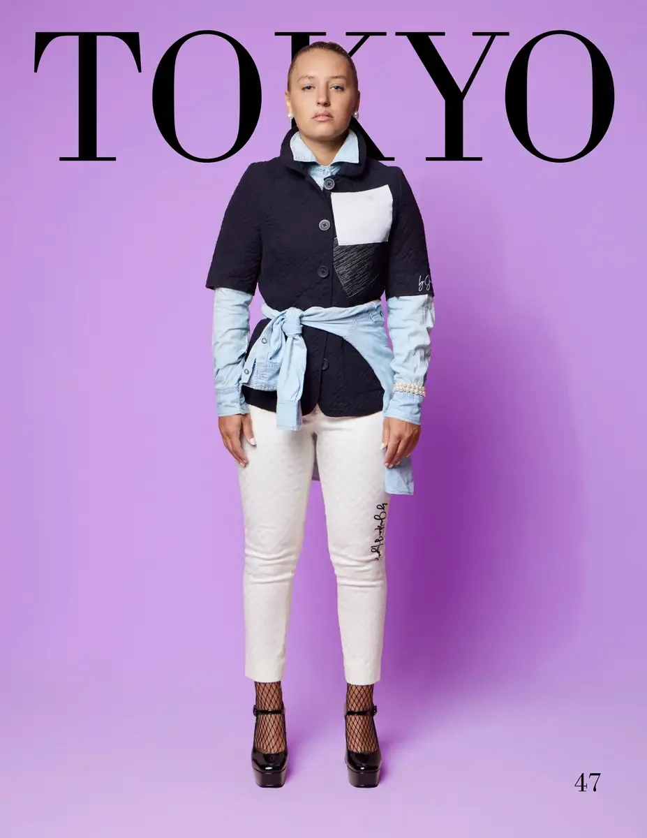 JUS10H 2025 Tokyo lookbook, a female model wearing a navy quilted short-sleeve jacket with white and black color-block patchwork panels and a script logo, layered over a light blue chambray shirt tied at the waist, cream skinny trousers with script e
