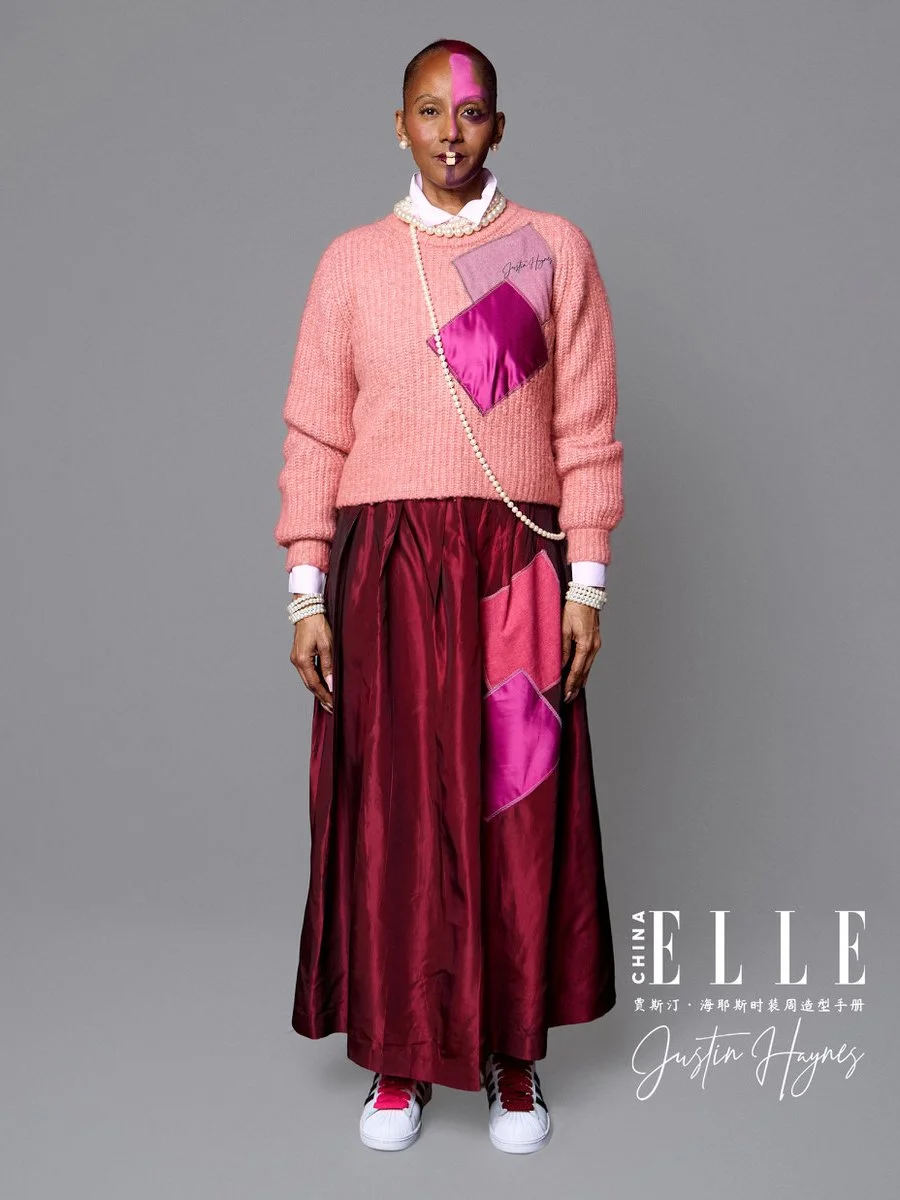 A woman wearing a pink sweater with a purple patch, a white collared shirt, pearl jewelry, and a long red satin skirt with pink patches, standing against a gray background.