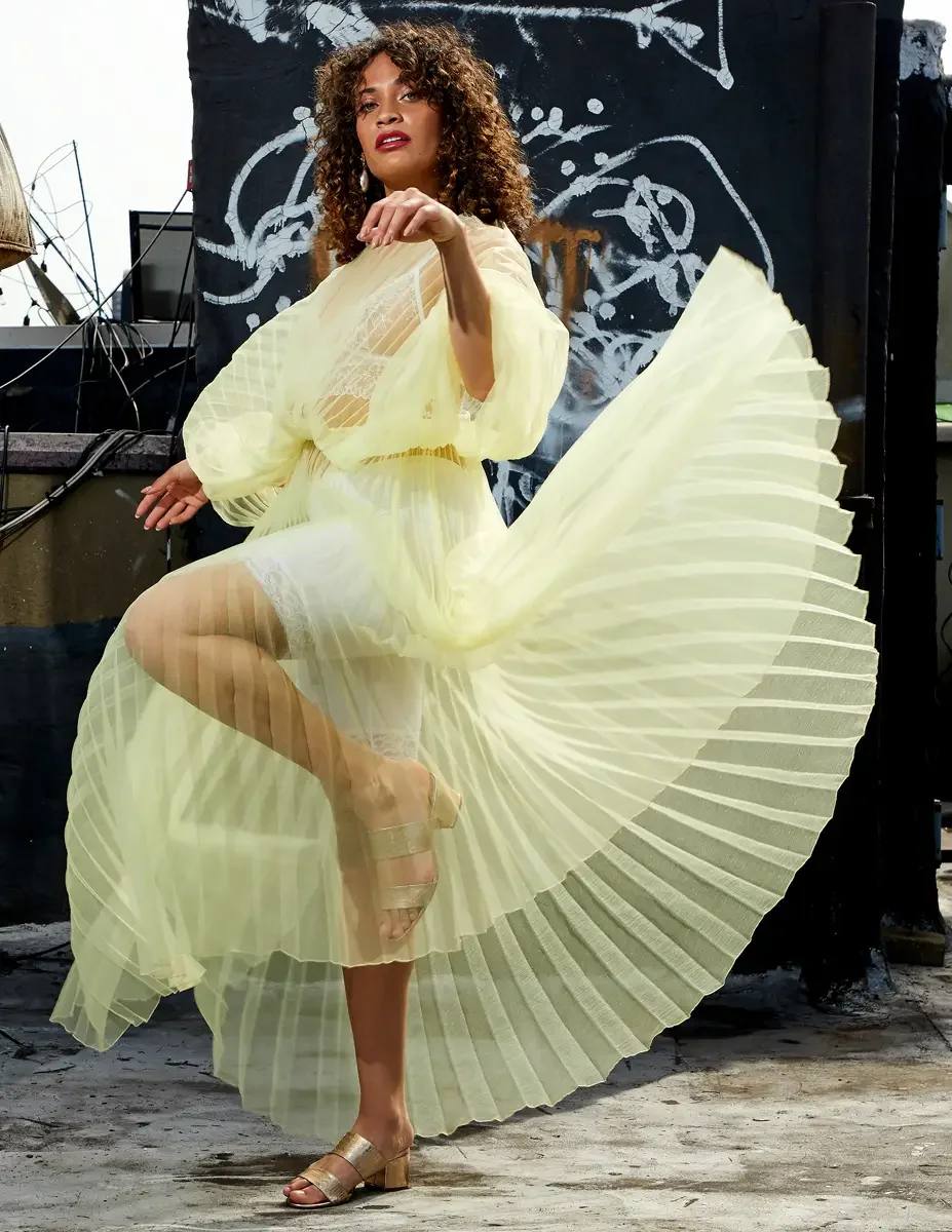 Fashion test shoot, full-length, model Ireland T in J'Amemme yellow pleated sheer gown, skirt lifted in motion, graffiti wall backdrop, East Village rooftop, New York City, styling by Matsine Stinson, makeup by Stefani Paige Vivian, photographed by S