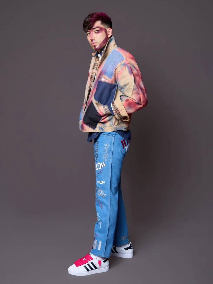 Young man standing sideways with hands in pockets, wearing a colorful mixed fabric jacket, blue jeans with text and graphics, and white sneakers with pink and black accents, against a plain gray background.