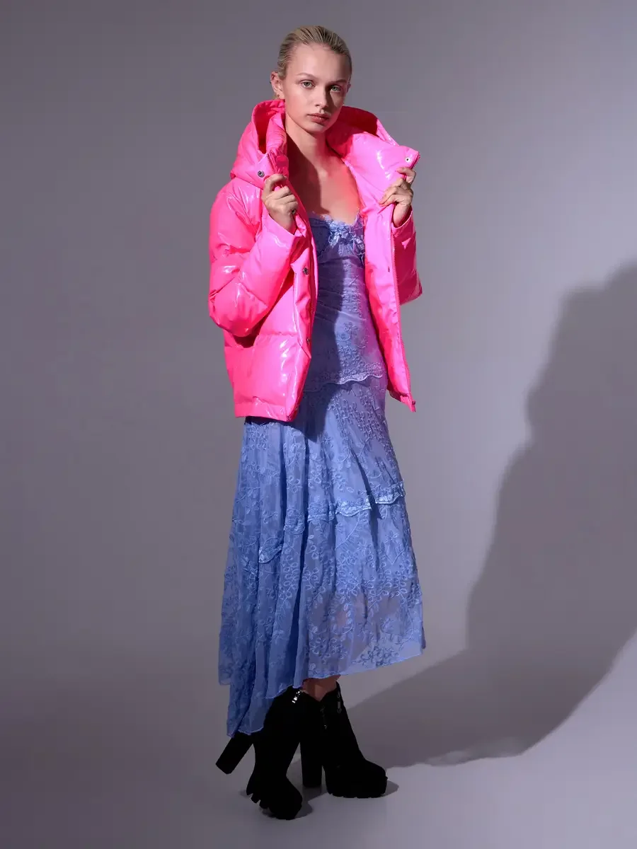 Model portfolio development session, high fashion editorial look, hot pink vinyl puffer jacket over periwinkle blue lace asymmetrical midi dress, full-length, slight turn to camera, both hands at collar, direct gaze, hard directional shadow defining 