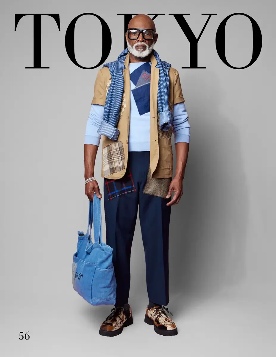 JUS10H 2025 Tokyo lookbook, a male model wearing a tan multi-pocket utility vest with mixed plaid, denim, and tweed patchwork appliqués, layered over a light blue crewneck sweatshirt and a blue and white striped shirt, navy wide-leg trousers, large b