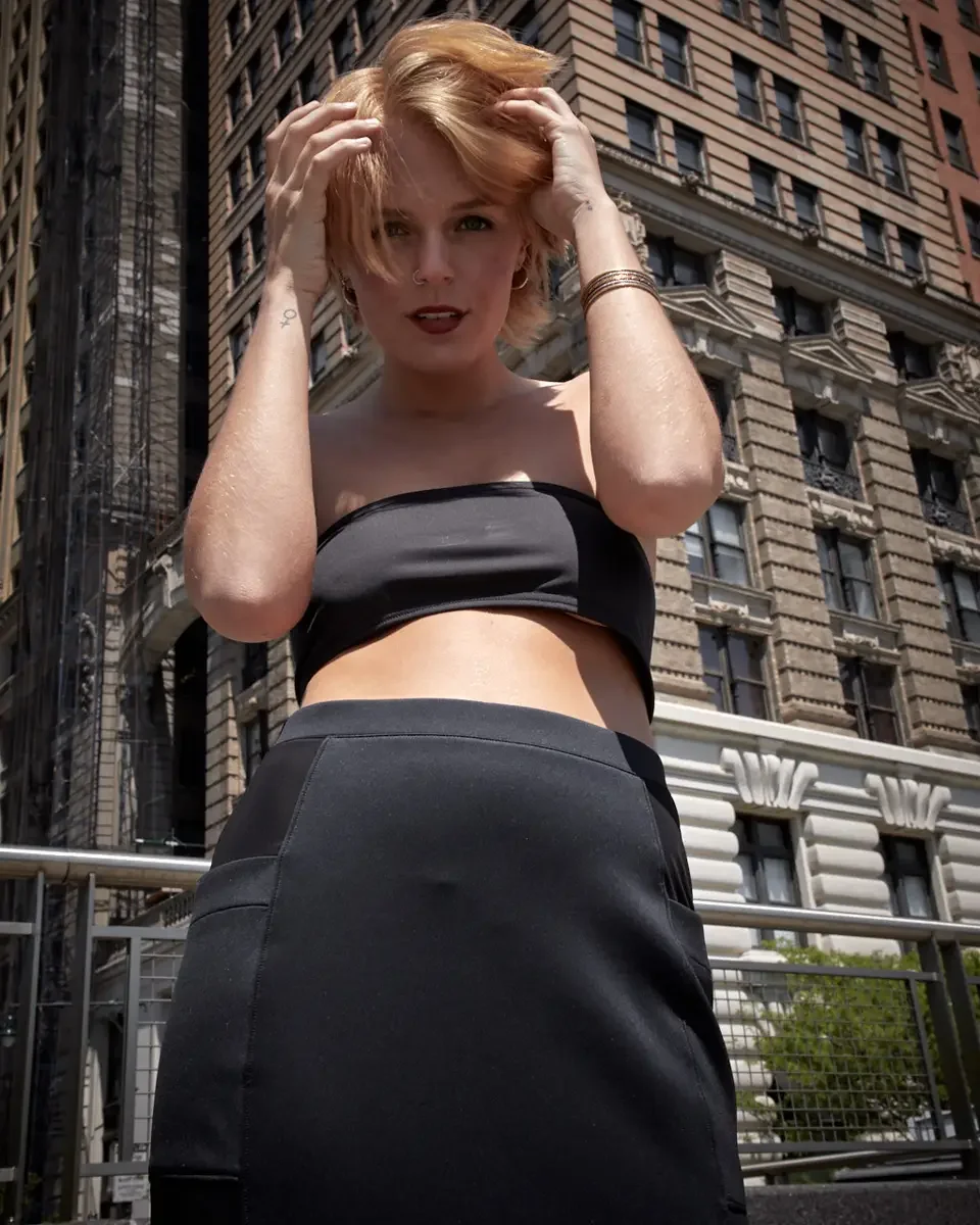 Satine, three-quarter crop, low camera angle, both hands in hair, black strapless bandeau crop top, black satin fitted midi skirt with wide waistband, gold bangles, small symbol tattoo on collarbone, nose ring, Beaux-Arts building in background, Batt