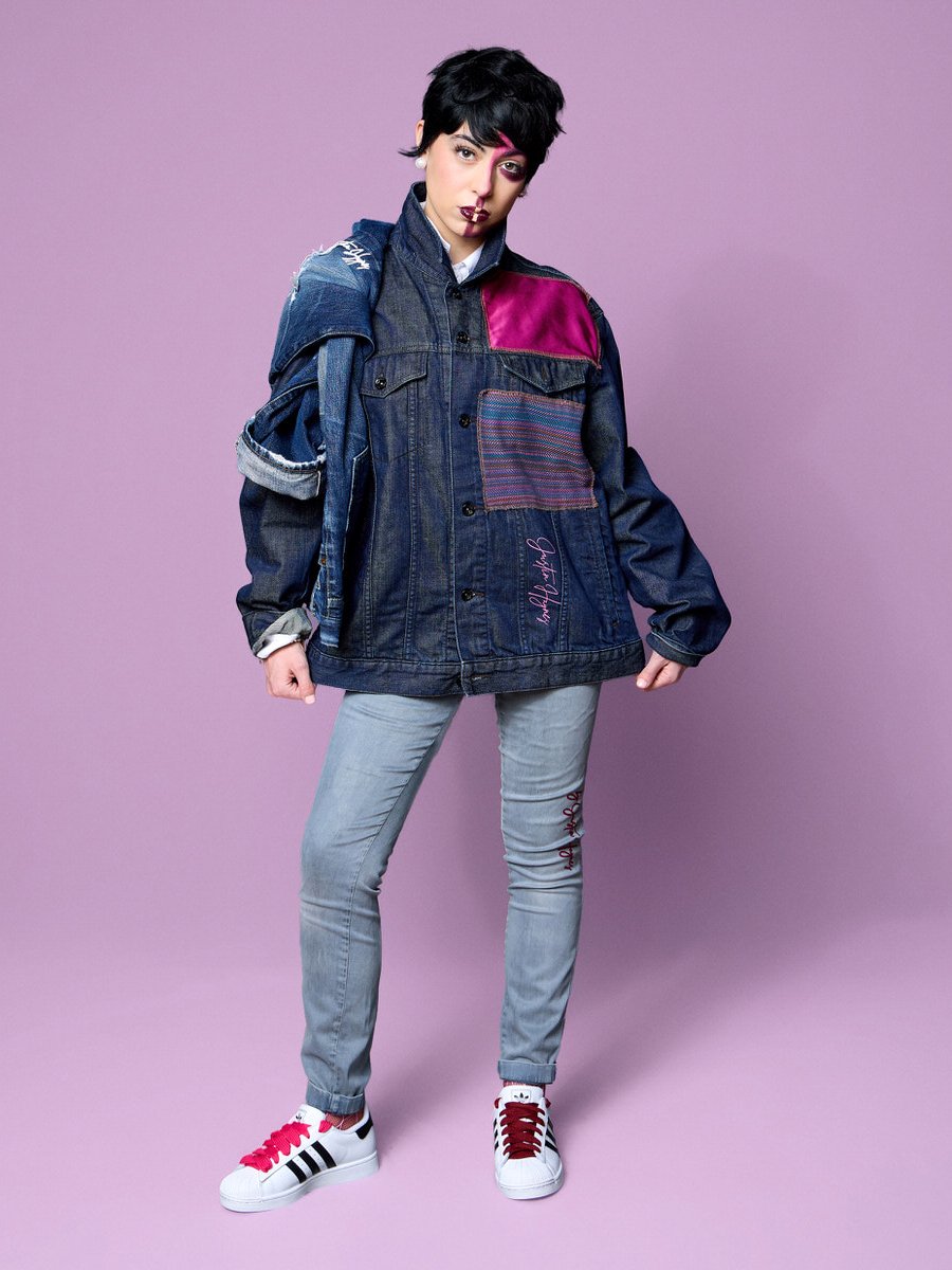 Young person with short black hair, wearing a layered denim jacket with pink and plaid accents, light gray jeans with embroidered text, and white sneakers with pink and black stripes, standing against a purple background.