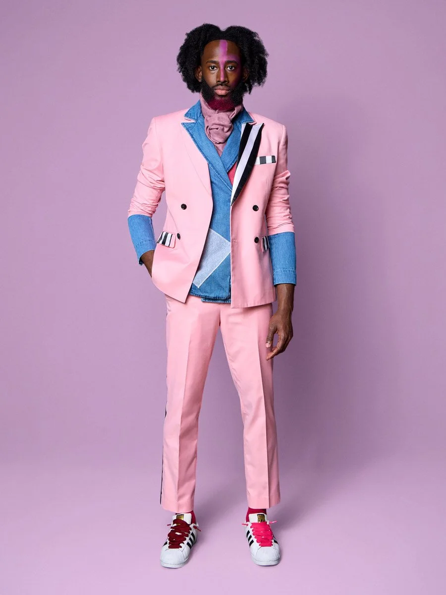 A man with dark skin and curly hair standing against a purple background, wearing a pink suit with blue and patterned accents, sneakers, and a pink scarf.