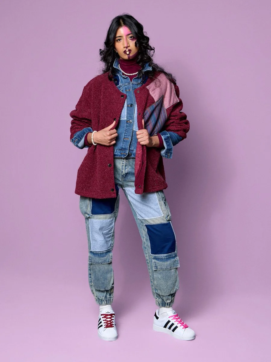 A young woman with dark curly hair wearing a layered outfit with a patchwork denim jacket, a maroon oversized cardigan, patchwork jeans, and white sneakers with black and pink accents, standing against a purple background.