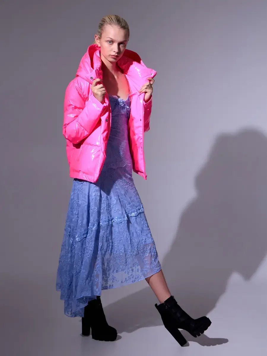 Model portfolio development session, high fashion editorial look, hot pink vinyl puffer jacket over periwinkle blue lace asymmetrical midi dress, full-length, three-quarter turn, both hands gripping lapels, weight shifted forward, directional shadow 