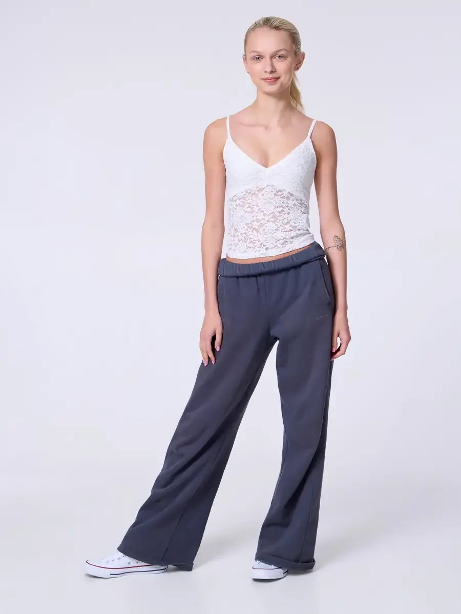 Model portfolio development session, commercial look, white lace crop camisole top and wide-leg charcoal sweatpants with elastic waistband, full-length, facing camera, weight shifted to one hip, easy smile, hands at sides, white Converse sneakers, li
