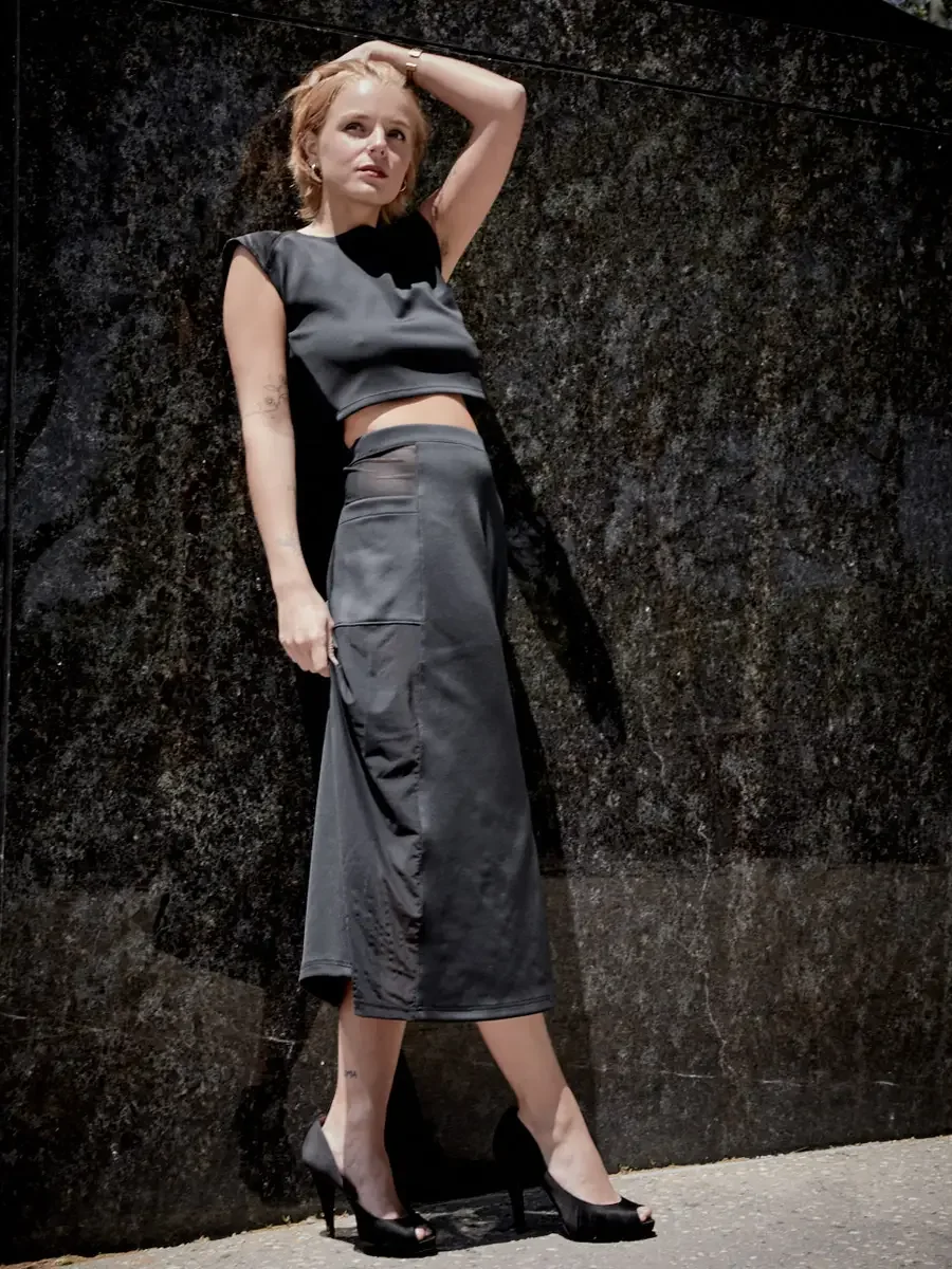 Satine, full length, one arm raised to head, black structured cap-sleeve crop top, charcoal grey A-line midi skirt with sheer mesh hip panel, black satin peep-toe stiletto heels, floral tattoo on left forearm visible, granite wall, Battery Park area 