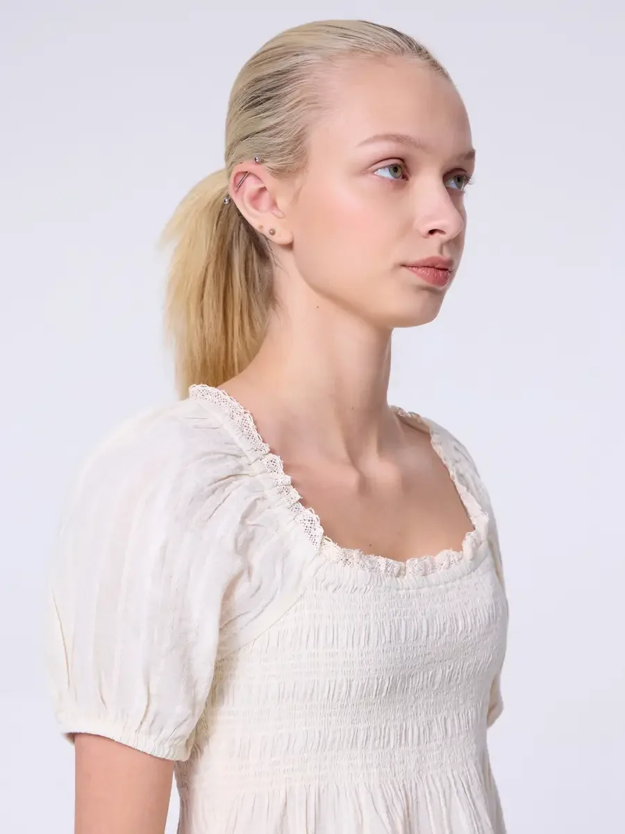 Model portfolio development session, commercial look, cream smocked square-neck puff-sleeve crop top with lace-trimmed neckline, three-quarter, profile turn, gaze directed off-camera, puff sleeve and smocked bodice shown in full relief, light gray ba