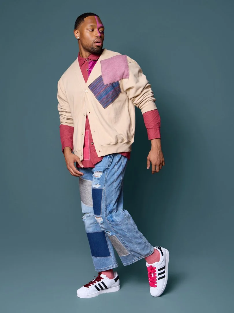 A young man in a beige patchwork jacket, red shirt, ripped jeans, and Adidas sneakers poses against a plain background.