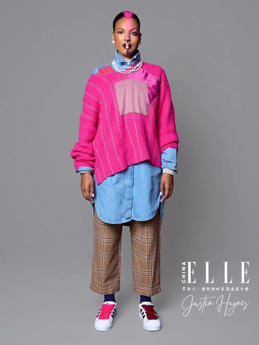 Fashionable woman wearing a pink oversized sweater layered over a blue denim shirt, paired with plaid pants and sneakers, with bold makeup and jewelry, on a gray background.