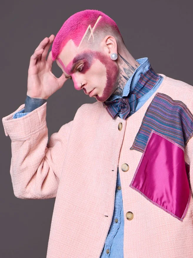 Person with bright pink hair, bold makeup, and a lightning bolt design on the head, wearing a light pink jacket with a pink pocket, layered over a blue denim shirt and patterned scarf.
