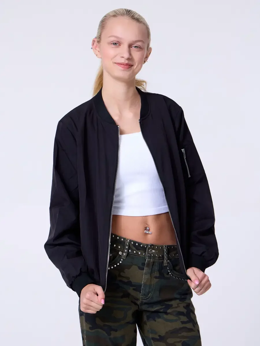 Model portfolio development session, commercial look, black bomber jacket open over white crop tank, low-rise studded camouflage wide-leg trousers with silver stud waistband detail, three-quarter, facing camera, hands loose at sides, easy smile, open
