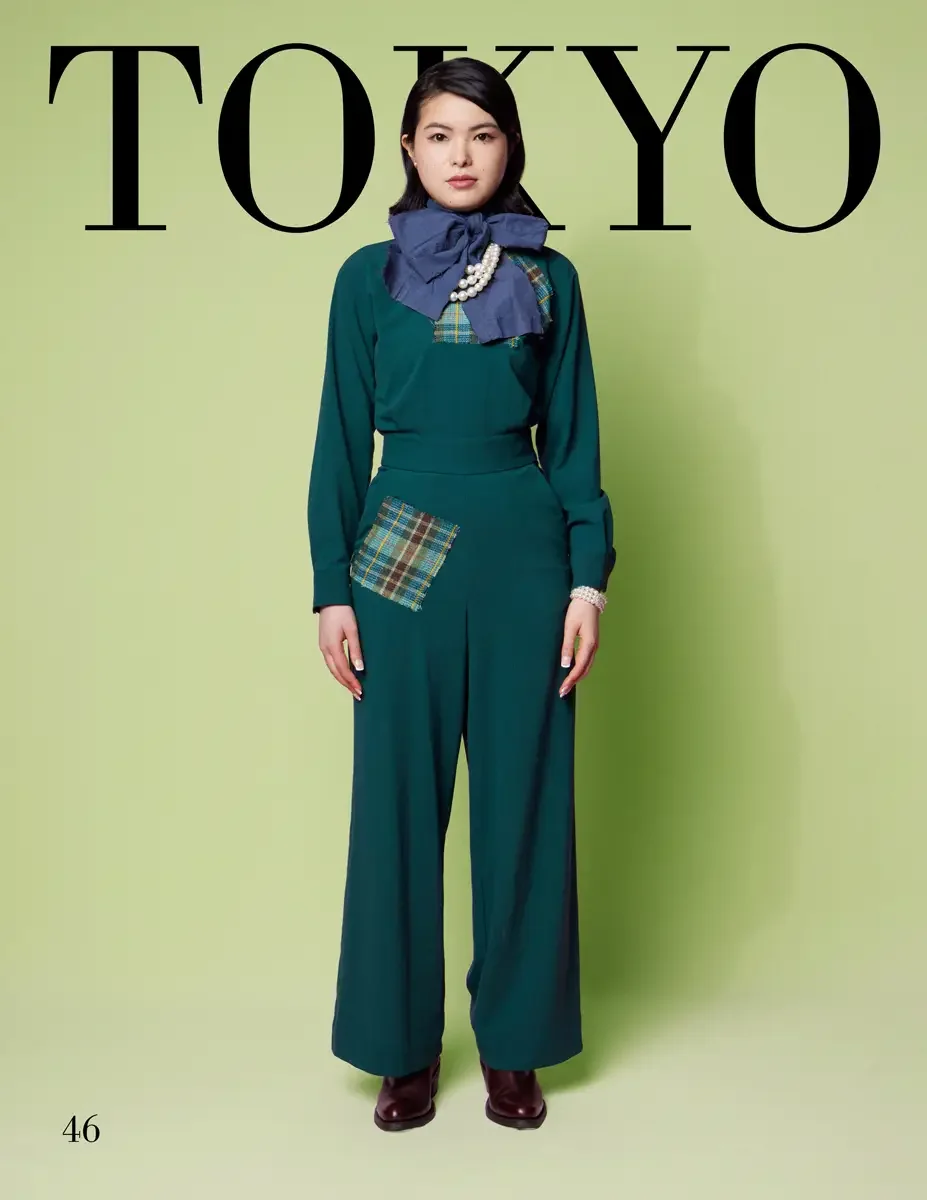 JUS10H 2025 Tokyo lookbook, a female model wearing a forest green long-sleeve jumpsuit with a tartan plaid patchwork pocket, a large slate blue denim bow at the neckline with a plaid accent, layered pearl necklaces, a pearl bracelet, and dark burgund