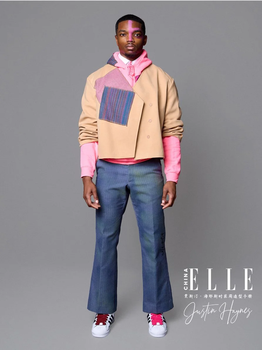 A young man in colorful layered clothing standing against a gray background, featuring a beige cropped jacket with patchwork details, pink hoodie, blue jeans, and sneakers.