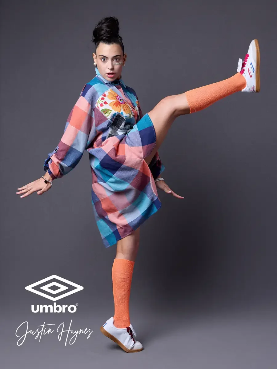 Editorial fashion photography for the Umbro France collection. Vanessa mid-kick with one leg raised high in an oversized multicolor plaid shirt dress with floral patchwork panels and orange football knee socks, styled by Justin Haynes, model Vanessa 