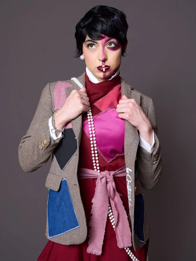A woman with short black hair and light skin wearing a beige blazer with blue and black patches, a maroon top, and a pink scarf, holding a long pearl necklace. Her face is painted with pink and purple makeup, and she stands against a gray background.