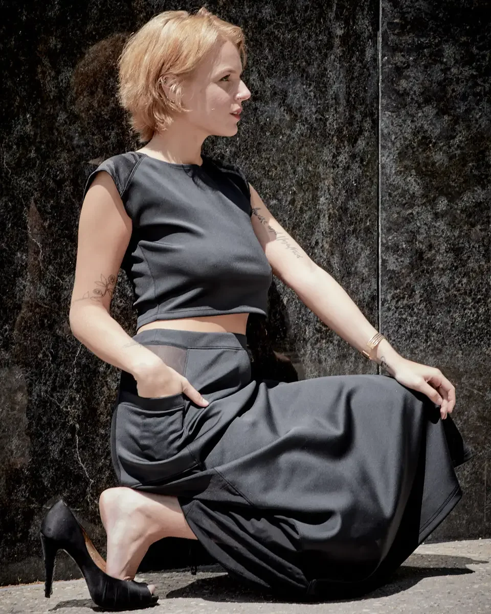 Satine, full length, seated on ground against granite wall, profile, hand in pocket, black structured cap-sleeve crop top, charcoal grey A-line midi skirt with sheer mesh hip panel, black satin peep-toe stiletto heels, gold bangle, floral tattoo on l