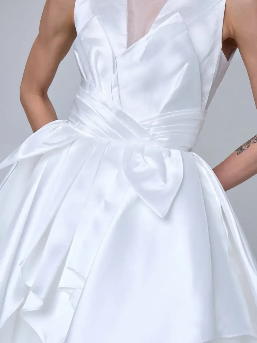 JUS10H 2025 New York Bridal Fashion Week, close detail of white sculptural satin ball gown with pleated V-neckline bodice, oversized structured bow at the waist, and cascading asymmetric peplum, photographed by Scott Parker Photo