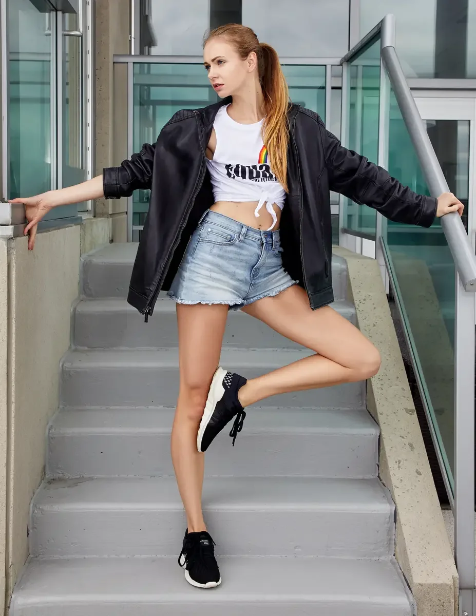 Julia, full length, standing on rooftop access stairs with one leg raised, white rainbow graphic tee knotted at the waist, light-wash frayed denim cutoff shorts, oversized black zip-front leather jacket, black chunky sneakers with white soles, Jersey