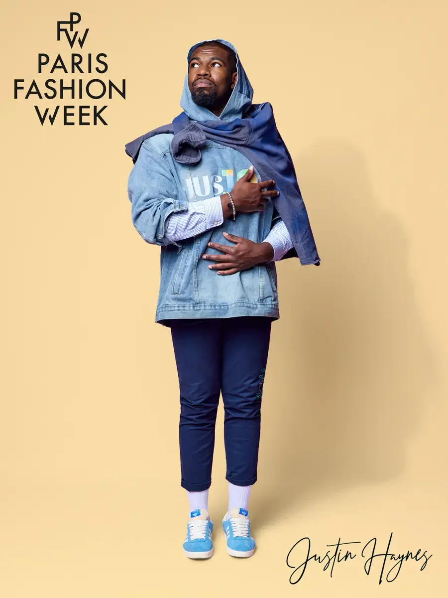 JUS10H 2025 promotional image, model standing with arms crossed, light wash JUS10H logo denim jacket layered over navy hoodie and striped shirt, navy JUS10H script trousers, white socks and sky blue sneakers, butter yellow background, photographed by