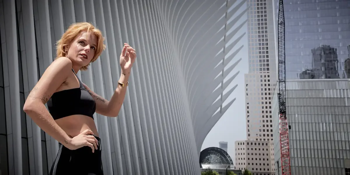 Satine, wide crop, body turned, one hand raised, black zip-front structured crop top, gold bangle, tattoos on both arms visible, Oculus World Trade Center, Battery Park area test shoot, photographed by Scott Parker Photo