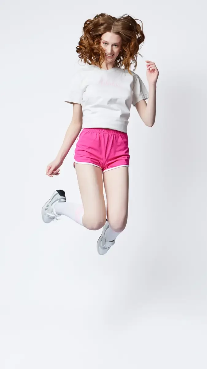Model portfolio development session, commercial athletic look, white Adidas tee, pink running shorts, white socks, silver sneakers, mid-jump pose, smiling, white background, photographed by Scott Parker Photo, New York