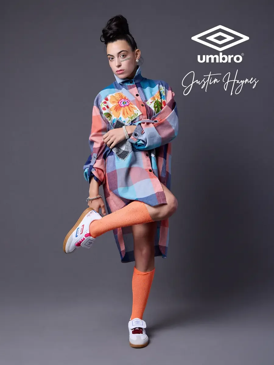 Elevated soccer style for the Umbro France collection. Vanessa balancing on one leg with knee raised in an oversized multicolor plaid shirt dress with floral patchwork panels and orange football knee socks, styled by Justin Haynes, photographed in Sp