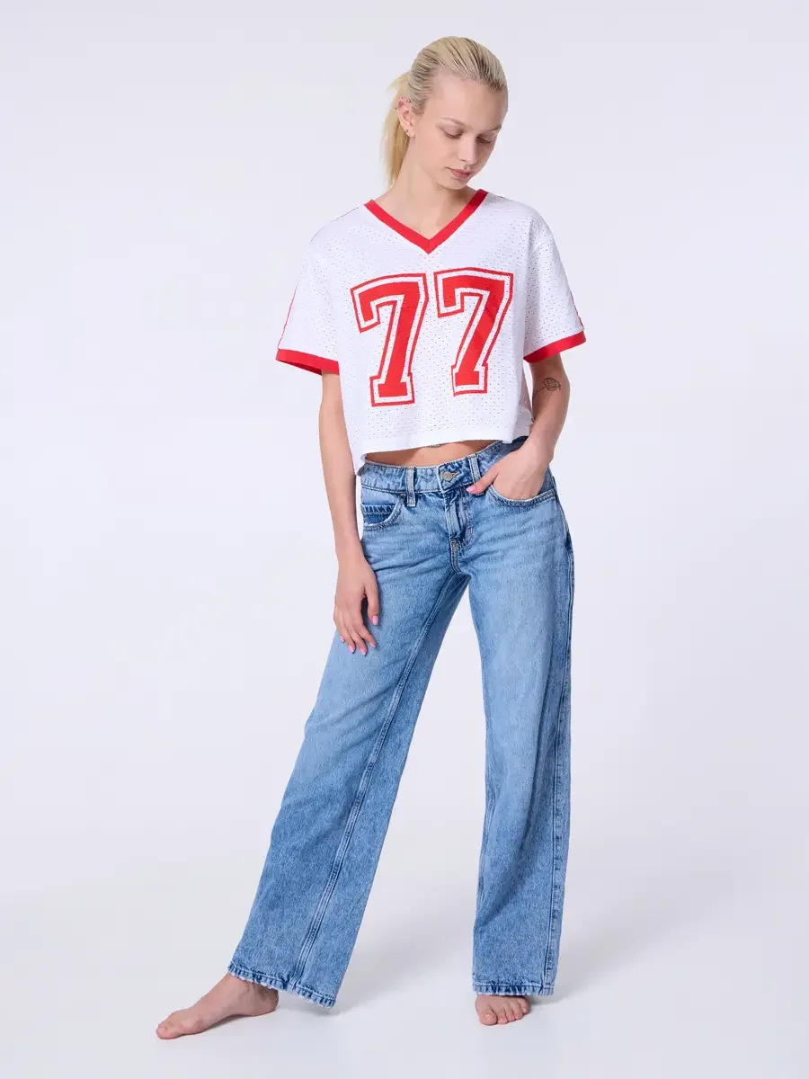 Model portfolio development session, commercial look, white mesh crop football jersey with red number 77 and red trim, low-rise light wash wide-leg jeans, full-length, slight turn, one hand in pocket, gaze downward, relaxed pose, jersey crop hem, bar