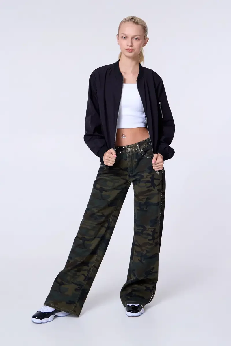 Model portfolio development session, commercial look, black bomber jacket open over white crop tank, low-rise studded camouflage wide-leg trousers with silver stud trim at waistband and side seam, full-length, facing camera, both hands at waistband, 
