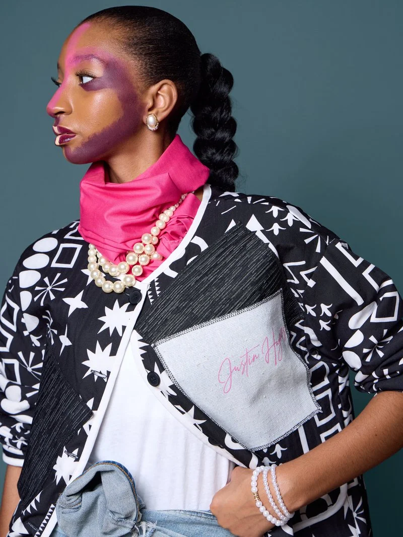 A woman with colorful makeup, black braided hair, wearing earrings, a pearl necklace, pink scarf, patterned black and white jacket, white shirt, gray jeans, and a pearl bracelet against a solid teal background.