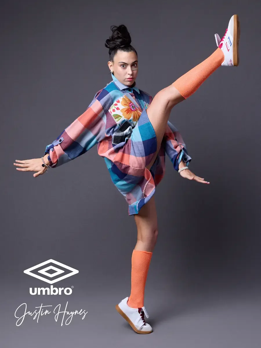 Fashion lifestyle photography for the Umbro France collection. Vanessa in a side profile high kick with arms extended for balance in an oversized multicolor plaid shirt dress with floral patchwork panels and orange football knee socks, styled by Just